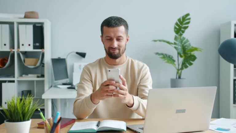 Mann smiler mens han ser pa smarttelefonen sin pa et moderne kontor
