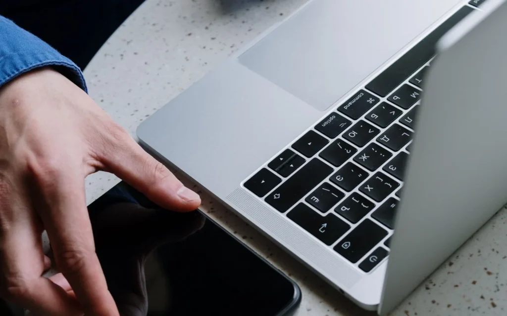Hånd på laptop med mobiltelefon liggende ved siden av på pulten