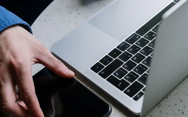 Hånd på laptop med mobiltelefon liggende ved siden av på pulten