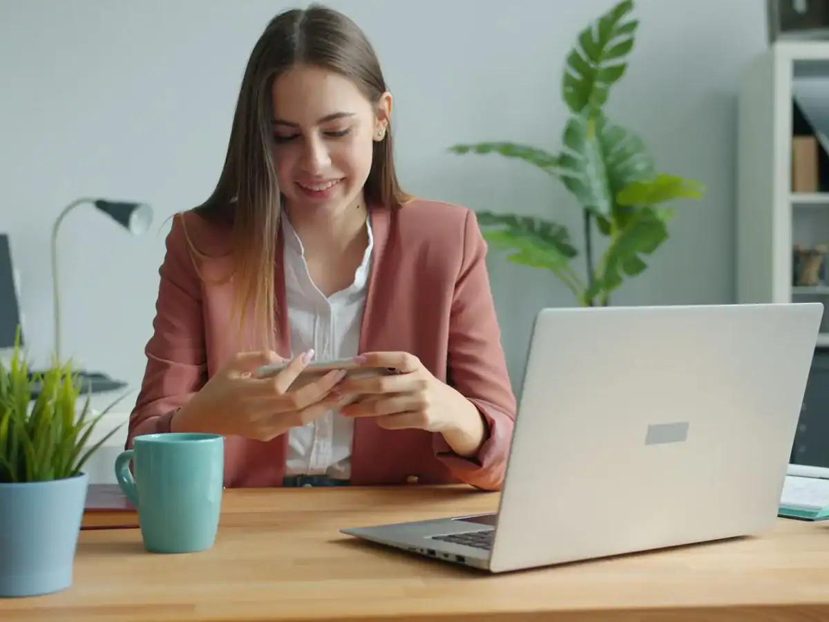 Ansatt i en liten bedrift sjekker mobilen ved laptopen på kontoret