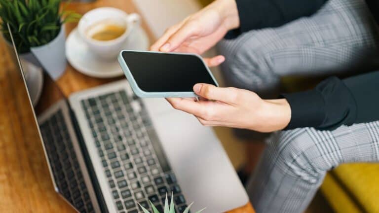 Hender som holder en smarttelefon over en bærbar PC på et skrivebord, med kaffekopp og planter i bakgrunnen